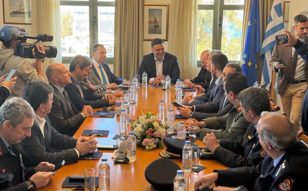 A formal meeting with officials seated around a long wooden conference table, water bottles and a floral centerpiece, while a camera crew films from the left and flags are visible in the background.